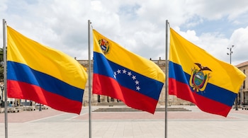 Tres banderas nacionales, Colombia, Venezuela (con escudo) y Ecuador (con escudo), ondean en mástiles frente a un edificio con columnas y un cielo nublado.