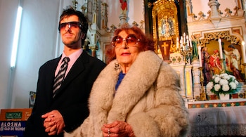 Un hombre joven con traje y gafas oscuras sostiene la mano de una mujer mayor con abrigo de piel y gafas frente a un altar con flores