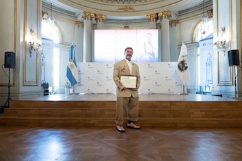 Adrián Appiolaza, vestido con un traje beige bordado, gorro rojo y zapatos rosas, sonríe en un escenario sosteniendo un certificado, con banderas y una pantalla detrás