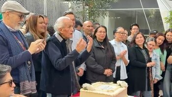 Hombre mayor de cabello blanco hablando, rodeado por varias personas, frente a una torta cuadrada con rosas blancas