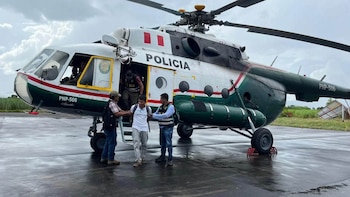 Un helicóptero blanco y verde de la policía estacionado en la pista mientras dos agentes guían a un hombre esposado