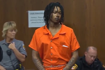 Un hombre joven con dreadlocks y un mono naranja de prisión, encadenado por la cintura, de pie en un tribunal flanqueado por dos agentes de seguridad