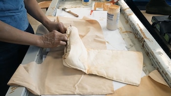 Manos trabajando en un zapato de cuero claro sin terminar, sujetando la piel con alfileres sobre una base de cuero beige en una superficie blanca. Un bote de pegamento es visible
