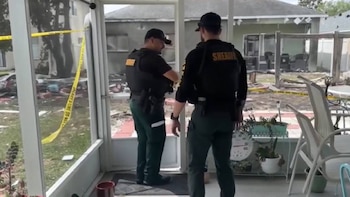 Dos agentes del Sheriff con uniforme oscuro y chalecos parados en una veranda. Se ve cinta policial amarilla "NO CRUZAR" y escombros en el fondo