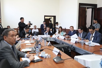 Personas en una reunión oficial alrededor de una mesa de conferencias con micrófonos y laptops. Un camarógrafo en el fondo documenta el evento