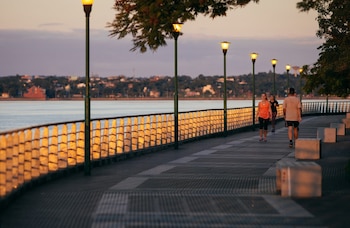 Vista de una costanera al atardecer con varias personas caminando. Hay farolas encendidas, una barandilla dorada por el sol y un cuerpo de agua con una ciudad al fondo