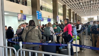 Vista frontal de pasajeros con equipaje haciendo fila en los mostradores de LATAM dentro de un aeropuerto, con barreras de control de acceso y señalización