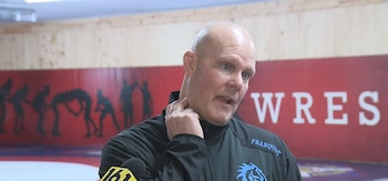 Un hombre calvo con chaqueta deportiva negra sostiene su mano en el cuello frente a un micrófono en una sala de lucha deportiva