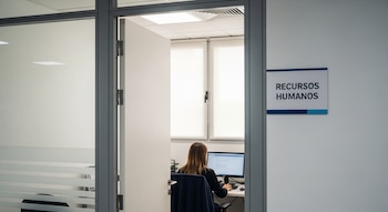 Una puerta de oficina abierta muestra a una mujer con cabello castaño sentada en un escritorio frente a una computadora. En la pared se lee Recursos Humanos.