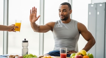 Hombre atlético de tez oscura y camiseta sin mangas gris rechaza un vaso de jugo de naranja. Frente a él, en una mesa, hay una botella de agua y frutas variadas.