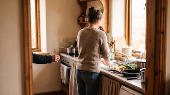 Mujer de espaldas en una cocina rústica, lavando en el fregadero. Una mano se extiende hacia ella. Verduras, ollas, sartenes y ventanas con luz.