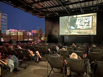 Vista trasera de una audiencia sentada en sillas reclinables en un cine al aire libre de Arthaus Central, viendo una película en una pantalla grande de noche