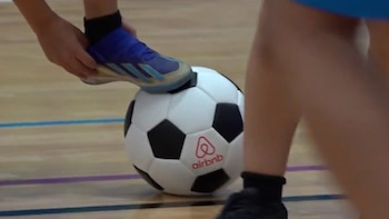 Un pie con zapatilla azul está sobre un balón de futsal blanco y negro con el logo de Airbnb en una cancha de madera con líneas de colores