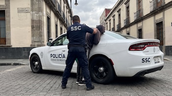 Un oficial de policía sostiene a una persona sobre un auto blanco en una calle empedrada frente a edificios antiguos.