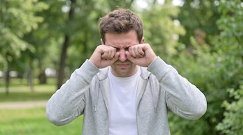 Un hombre con chaqueta gris y camiseta blanca se frota los ojos con los puños en un espacio verde con árboles al fondo.