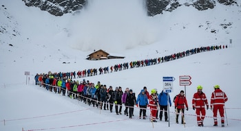 Larga fila de personas camina por ruta nevada en montaña, guiadas por rescatistas. Un edificio de madera y una masa de nieve descendida se ven al fondo.