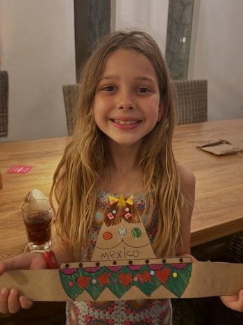 Una niña sonriente sentada ante una mesa de madera mientras sostiene con ambas manos un adorno de cartón con forma de sombrero y la palabra México