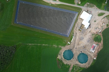 Vista aérea de una granja con un estanque rectangular, dos tanques circulares, edificios blancos, áreas de tierra y campos verdes