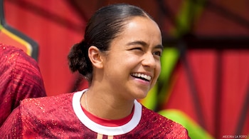 Una mujer sonríe y guiña un ojo con el cabello recogido en un moño y vistiendo una camiseta roja con detalles dorados y cuello blanco.