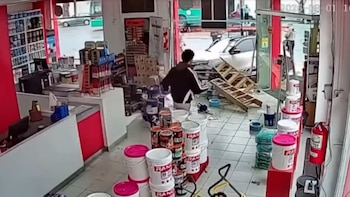 Interior de una pinturería con un automóvil plateado que ha atravesado la entrada, daños materiales, un hombre, latas de pintura y una escalera rota