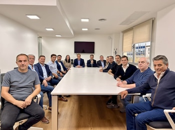 Un grupo de trece personas, en su mayoría hombres, sentados alrededor de una larga mesa blanca en una sala de reuniones con una pantalla y una ventana