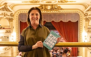 Retrato de Alejandra Naughton, una mujer con cabello castaño, sonriendo y sosteniendo un libro verde, en un balcón de teatro ornamentado