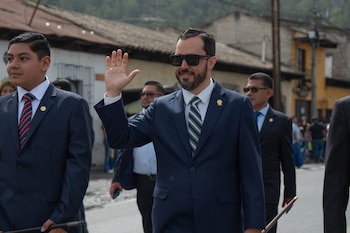Un hombre con traje azul, gafas de sol y corbata a rayas camina por una calle con la mano derecha levantada junto a personas en traje