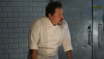 Retrato de Jon Favreau vestido con una chaqueta de chef blanca, con tatuajes visibles en los brazos, en una cocina con azulejos y una puerta metálica