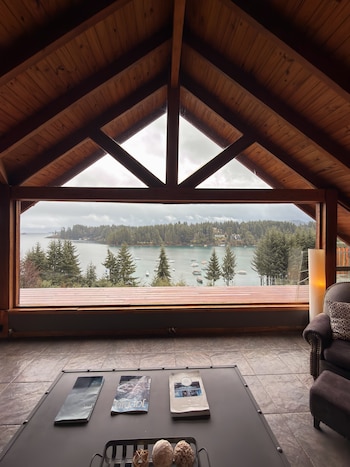 Interior de una cabaña de madera con un gran ventanal frente a un lago con barcos, una mesa de centro con revistas y un sillón