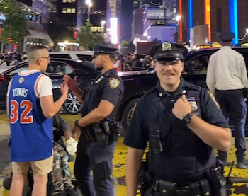 Dos policías interactúan con un aficionado que lleva una camiseta azul con "Towns 32" en una calle de noche, con vehículos y edificios iluminados