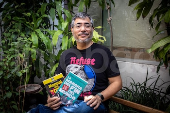 Hombre con gafas y barba canosa sentado, sostiene dos libros. Viste camiseta negra con estampado y jeans. Fondo con plantas y pared