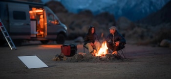 Una antena rectangular en el suelo, dos personas junto a una fogata y una furgoneta iluminada en un paisaje montañoso al anochecer