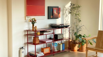 No canto da sala com estante vermelha, toca-discos, dois alto-falantes, livros, plantas e quadros na parede, com luz solar direta.