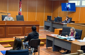Un tribunal con un hombre en estrado, dos mujeres en escritorios, un hombre de espaldas y otro en pantalla de videoconferencia. Documentos y micrófonos