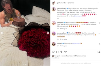 Isaac Moreno sonriendo, sentado en la cama, sostiene un pastel de chocolate con una vela. A su lado, un enorme ramo de rosas rojas envuelto en papel negro