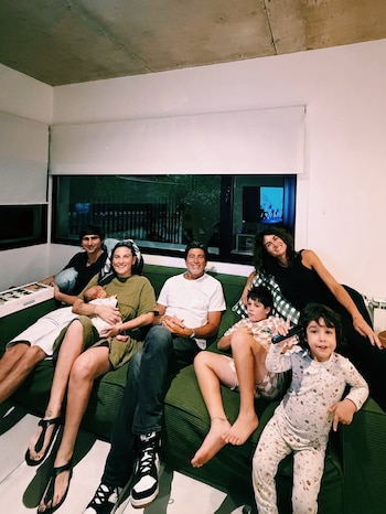 Una familia numerosa de siete personas, incluyendo un bebé, sentada en un sofá verde. Se observa a adultos y niños sonriendo en un ambiente hogareño