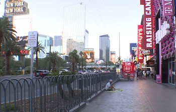 Acera con peatones, valla metálica, palmeras, calle con vehículos, edificios altos y carteles luminosos de comercios en una avenida