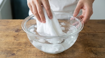 Manos sumergiendo una toalla blanca en un recipiente de cristal transparente lleno de agua y cubos de hielo, sobre una mesa de madera.