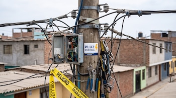 Un poste de madera con cables, una caja de conexiones abierta, herramientas colgadas, cinta amarilla de peligro y un logo de Pluz Energía.