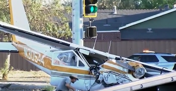 Avioneta blanca y amarilla con el frente dañado junto a un poste con semáforo, un hombre cerca de la cabina y una patrulla al fondo