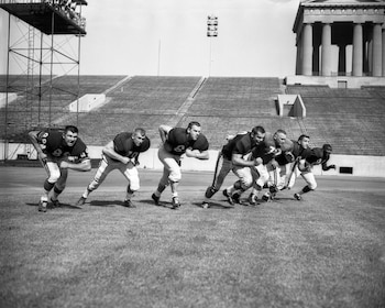 Siete jugadores de fútbol americano en postura de carrera sobre césped dentro de un estadio vacío con gradas y columnas al fondo