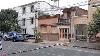 Calle con una camioneta policial azul y casas de fachada clara, acordonada con cinta de seguridad roja y blanca por un operativo policial