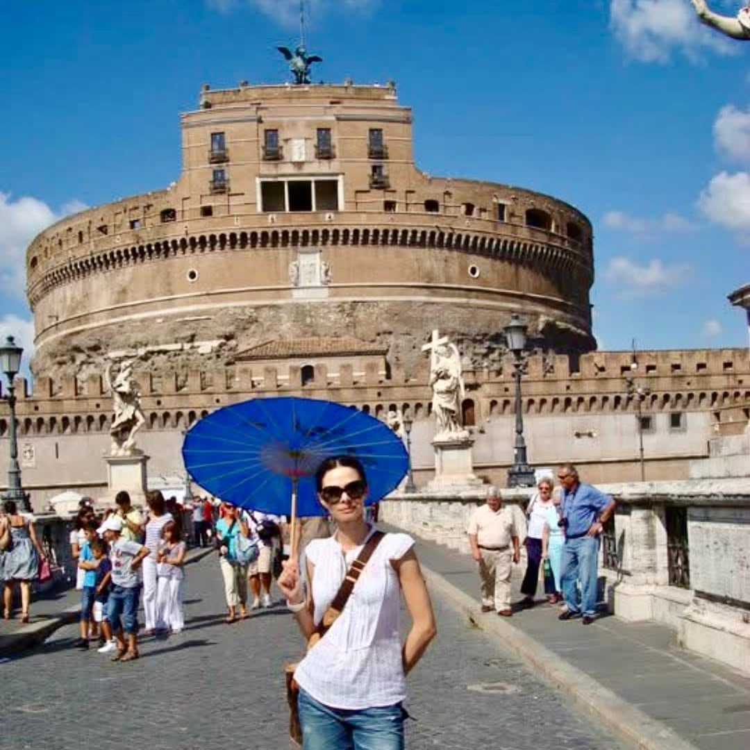 La exmodelo posando con un parasol azul frente al icónico Castillo Sant'Angelo de Roma en un día soleado, durante un viaje a la capital italiana