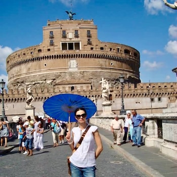 Mujer con gafas de sol y parasol azul posa en un puente. Detrás, el Castillo Sant'Angelo y estatuas bajo un cielo azul. Varias personas en el fondo