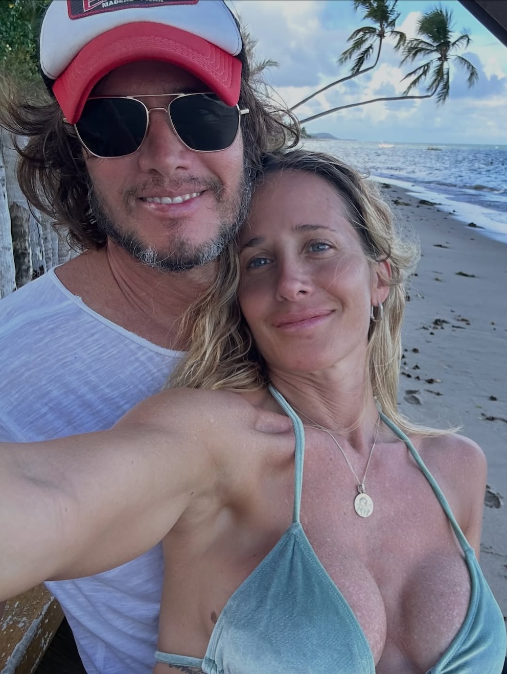 Julieta Prandi y Emanuel Ortega sonríen frente al mar; él lleva gorra roja y gafas de sol, y ella, bikini celeste