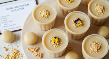 Varias mini gelatinas de mazapán en vasos, decoradas con cacahuetes triturados y flores, sobre una tabla de madera. Bolas de mazapán y cacahuetes a la vista.