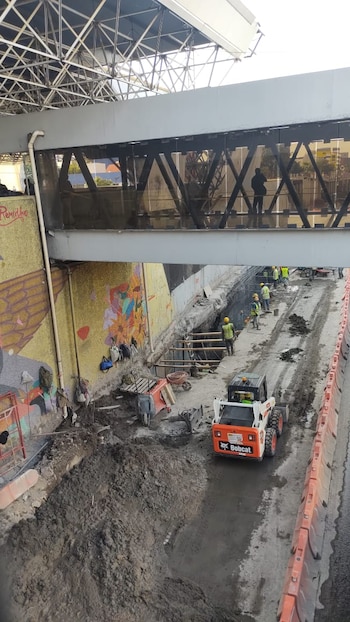 Obreros con chalecos amarillos y cascos realizan trabajos de excavación con una minicargadora Bobcat en una zanja, bajo una pasarela elevada