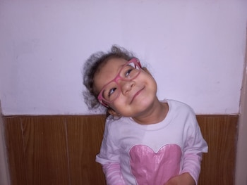 Retrato de Emma Rodriguez, una niña con gafas rosadas y camiseta blanca con corazón rosa, sonriendo en un fondo de pared blanca y madera