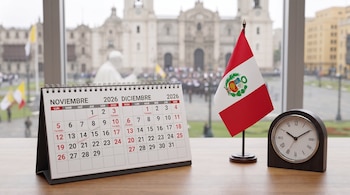 Un calendario de escritorio de 2026, una bandera de Perú y un reloj analógico sobre una mesa frente a una ventana.