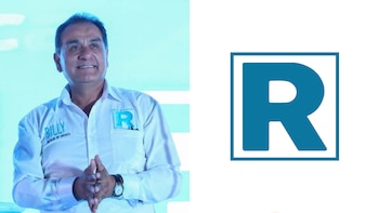Un hombre viste una camisa blanca con las manos juntas frente a un panel blanco con el logotipo de la letra R azul